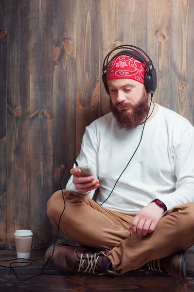 轻松的年轻胡子男子在耳机与智能手机坐在靠近木墙和听音乐 — 图库照片
