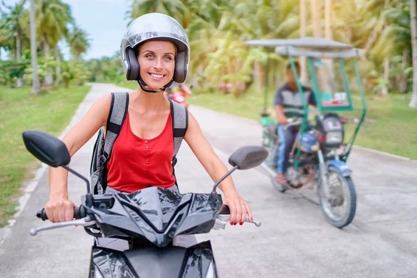 Happy young woman in helmet riding scooter on the road with palm trees.