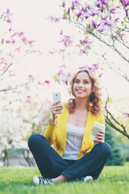 Bahar park çimenlerin üzerinde otururken kulaklık Smartphone kullanarak çekici genç kadın.