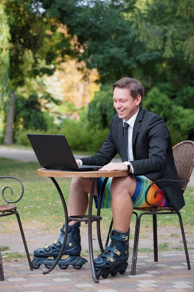 Businessman dressed in suit, shorts and rollers working with laptop at the outdoors cafe