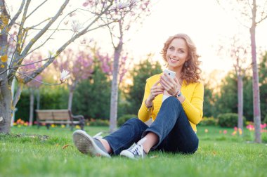 Bahar park çimenlerin üzerinde otururken Smartphone kullanarak çekici genç kadın.