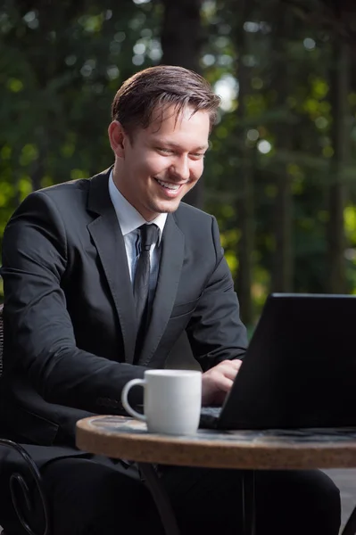 Attractive young caucasian man in formal wear working on laptop and smiling while sitting at the table outdoors.