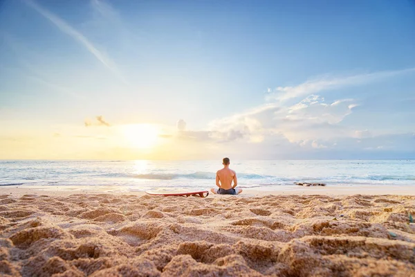 放松的年轻人坐在莲花位置与冲浪板在海滩上 — 图库照片