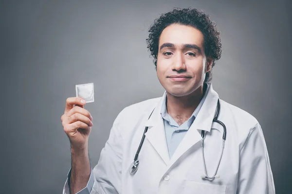 Handsome arabic doctor in medical uniform holding condom while standing against grey background.