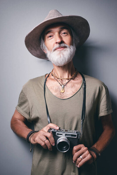Studio portrait of handsome senior man with gray beard and hat holding photo camera.
