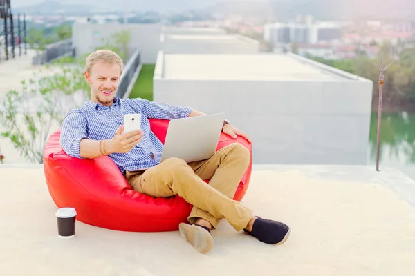 年轻英俊的人在笔记本电脑上工作 并使用智能手机 而坐在红色的豆子袋 — 图库照片