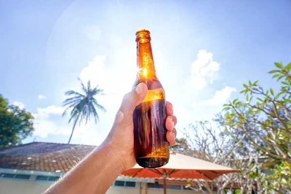 Male hand holding bottle of beer against sunny sky.