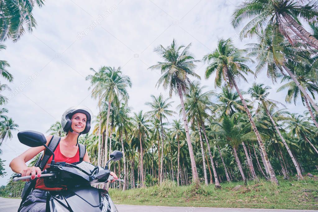 Young beautiful woman in helmet riding scooter on the road with palm trees.