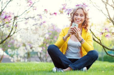 Bahar park çimenlerin üzerinde otururken kulaklık Smartphone kullanarak çekici genç kadın.