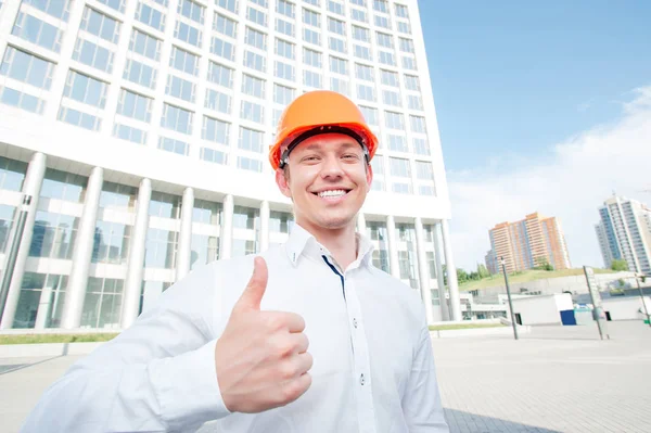 Cheerful young man in shirt and hardhat showing thumb up and smiling while standing outdoors