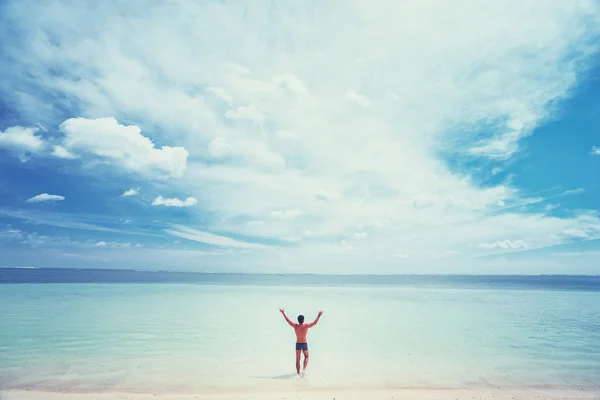 Happy young man rising hands up standing on tropical beach enjoying beautiful view.