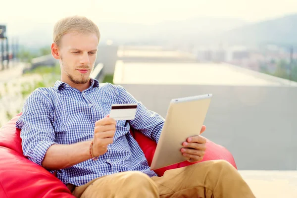 赤のビーンバッグに座ってクレジット カードを保持しているタブレット コンピューターを使用してハンサムな若い男 — ストック写真