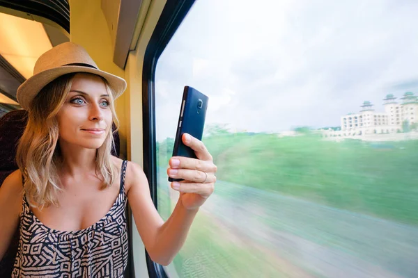 若い可愛い女性がスマート フォンを使用してウィンドウのそばに座って電車旅行写真を撮る — ストック写真