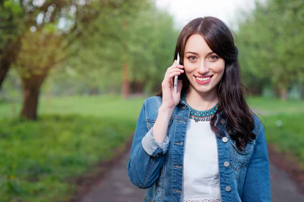 Attractive young woman talking by smartphone while walking by summer park.