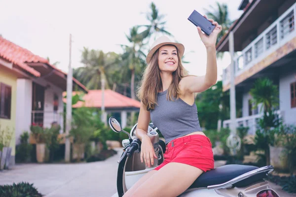 彼女のスマート フォン Selfie を取ってスクーターの上に座って帽子でかなり若い女性の屋外のポートレート — ストック写真