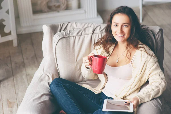 Beautiful young woman with cup of tea using tablet computer while sitting on armchair at home.