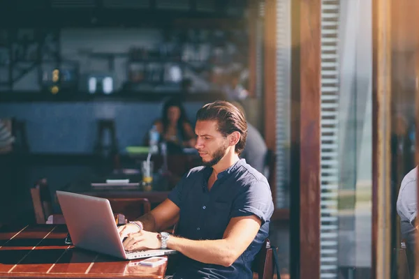 坐在咖啡厅露台的年轻胡子男子在笔记本电脑上工作 — 图库照片