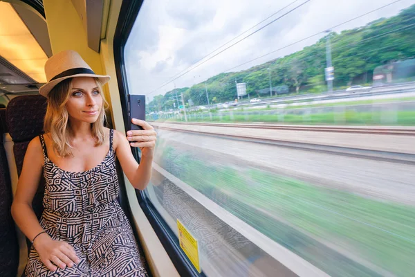 若い可愛い女性がスマート フォンを使用してウィンドウのそばに座って電車旅行写真を撮る — ストック写真