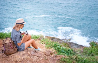 Deniz manzaralı cliff üzerinde oturan kamera tutarak sırt çantası ile şapkalı genç kadın.