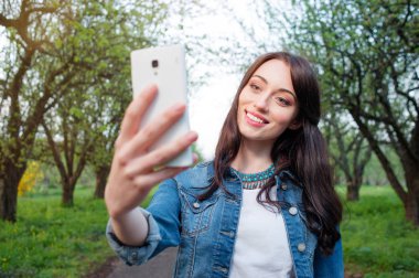 Yanında yeşil park smartphone selfie alarak genç gülümseyen kadın.