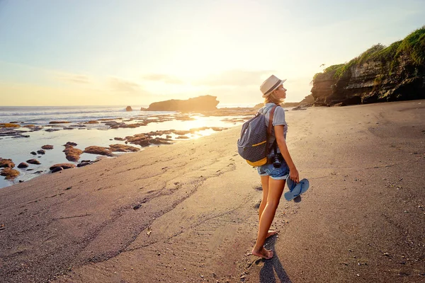 カメラとバックパックの夕日を楽しむ海のビーチで裸足で歩く若い女性 — ストック写真