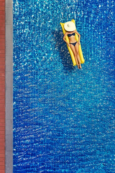 Top view of slim young woman in bikini on the yellow air mattress in the big swimming pool.