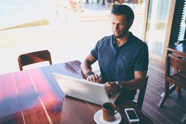 坐在咖啡厅露台的年轻胡子男子在笔记本电脑上工作 — 图库照片