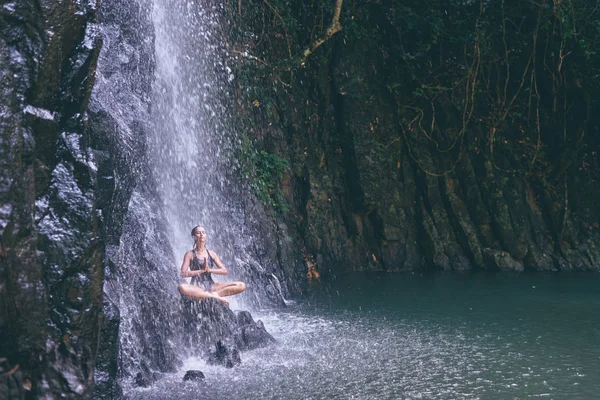 穿着泳装的漂亮年轻女子坐在热带瀑布下的岩石莲花位置上 — 图库照片