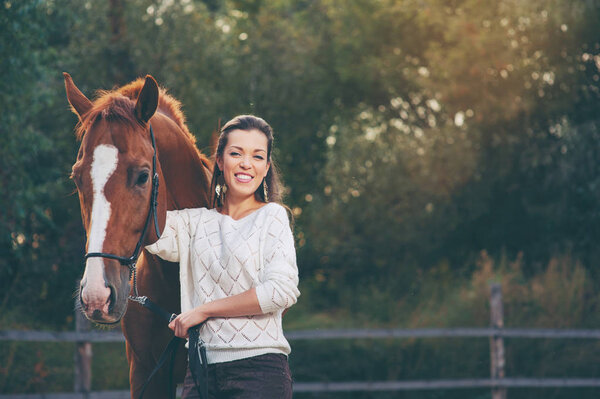 Young smiling beautiful woman walking with brown horse outdoors