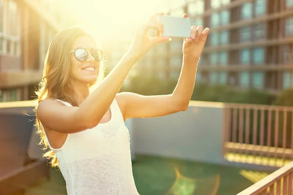 かなり若い女性サングラス都市背景に彼女のスマート フォンのカメラで写真を撮る — ストック写真