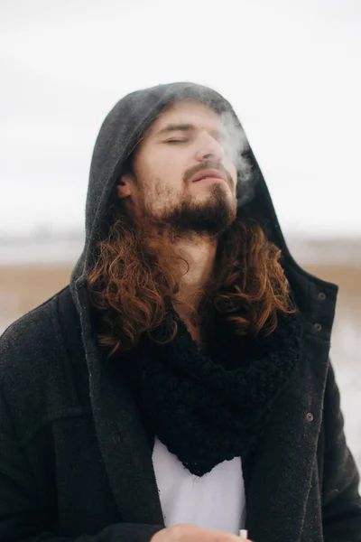 Seriously bearded man with long hair smoking in the field. Wearing black coat.