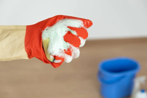 Manos en guantes para limpiar. Una esponja con un detergente se espumó en una mano sobre un cubo azul. — Foto de Stock