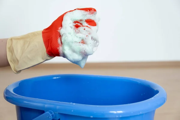 Manos en guantes para limpiar. Una esponja con un detergente se espumó en una mano sobre un cubo azul. — Foto de Stock