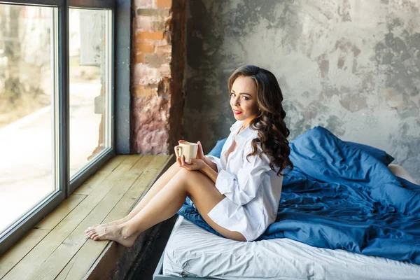 Petite brunette caucasian girl enjoing morning coffee in bed in loft room