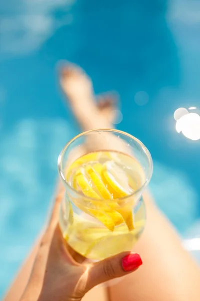 female hand holding glass of water with lemon over swimming pool