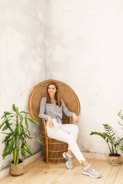 Genç güzel kız beyaz pantolon ve mavi gömlek beyaz oda ahşap bir sandalyede oturuyor