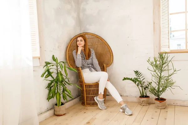 Genç güzel kız beyaz pantolon ve mavi gömlek beyaz oda ahşap bir sandalyede oturuyor