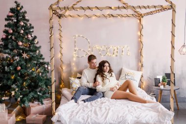 Genç güzel çift yatakta Noel ağacı arka plan üzerinde oturan ve gülümseyen