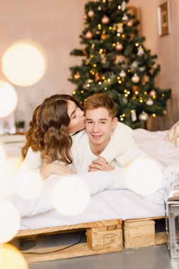 Genç güzel çift yatakta Noel ağacı arka plan üzerinde oturan ve gülümseyen