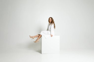 young beautiful girl in white clothes sitting in a white room on a white cube