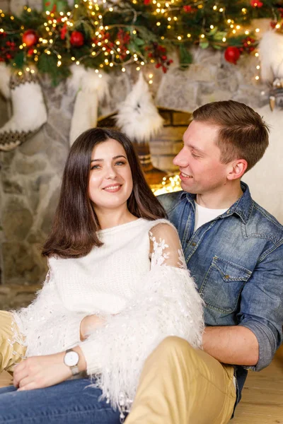 Young beautiful couple is sitting near the fireplace on the background of the Christmas tree and smiling