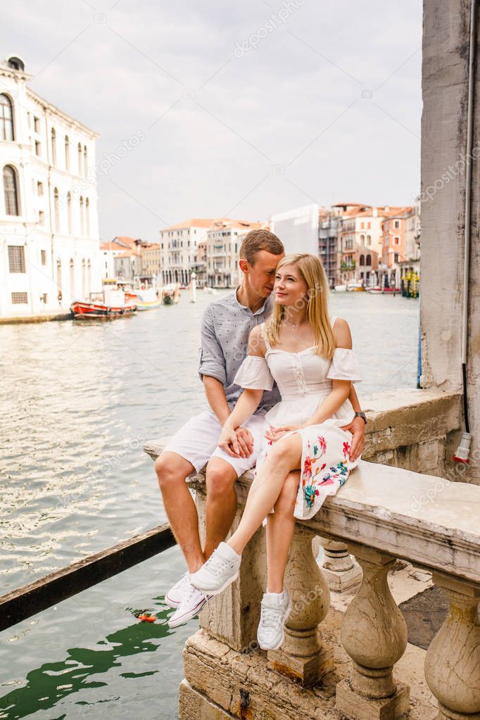 Young beautiful couple, girl in white dress and man in blue shirt are sitting on the balcony in the beautiful city of Venice in Italy