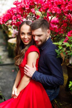 Kırmızı elbiseli genç ve güzel bir çift. Mavi gömlekli bir adam. Bahçede çiçekli duruyor.