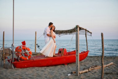 beyaz giysiler içinde genç güzel çift gün batımında denize yakın plajda kırmızı bir tekne üzerinde duruyor