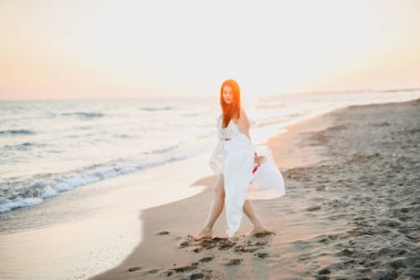 beyaz elbiseli genç güzel kız gün batımında denize yakın sahilde yürüyor