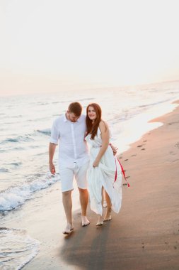gün batımında denize yakın sahilde yürüyen beyaz giysiler içinde genç güzel çift