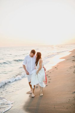 gün batımında denize yakın sahilde yürüyen beyaz giysiler içinde genç güzel çift