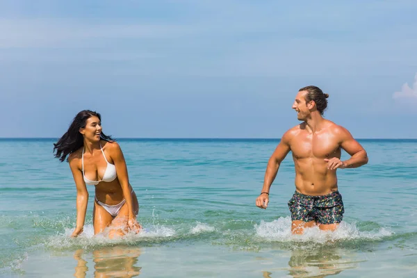Happy couple laughing together holding hands running having fun splashing water in the ocean waves. Young beautiful fit slim body people enjoying their happy lifestyle in paradise destination beach.