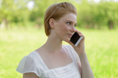 Gülümseyen güzel genç bir kadının portresi cep telefonuyla yaz yeşili parkın arka planına yakın..