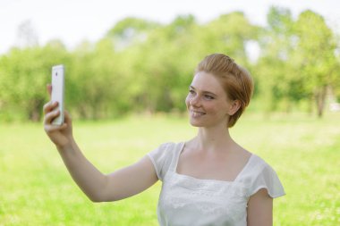 Gülümseyen güzel genç bir kadının portresi cep telefonuyla yaz yeşili parkın arka planına yakın..